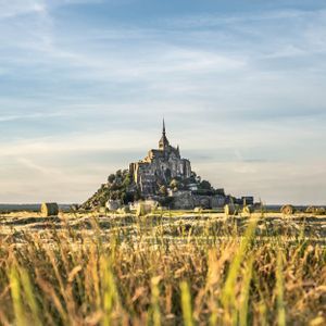 Eine historische Burg auf einem felsigen Hügel, gesehen über ein Feld voller hohem Gras und Heuballen im Sonnenuntergang.