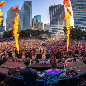 Una vista da dietro due DJ sul palco che guardano una folla immensa a un festival, con fiamme pirotecniche e grattacieli.