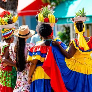 Quatre femmes vues de dos, vêtues de robes traditionnelles éclatantes, équilibrant des paniers de fruits sur leur tête en marchant.