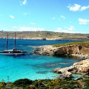 Un barco de dos mástiles está anclado en una cala rocosa con aguas claras y turquesas bajo un cielo azul.