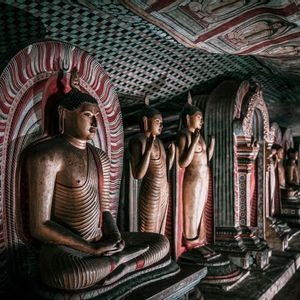 Una lunga fila di statue di Buddha scolpite all'interno di un tempio in grotta poco illuminato, con un soffitto riccamente decorato.