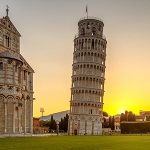Ein schiefer Glockenturm und eine kunstvolle Kathedrale stehen auf einem grünen Rasen vor einem goldenen Sonnenuntergang.