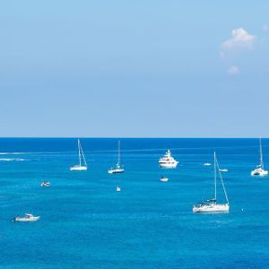 Vista panoramica di diverse barche a vela su un mare blu intenso, accanto a una grande scogliera rocciosa, sotto un cielo limpido.