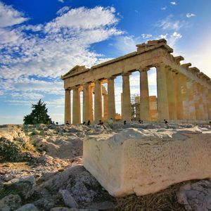 An ancient Greek temple with large columns stands on a rocky hill under a blue sky with white clouds at sunset.