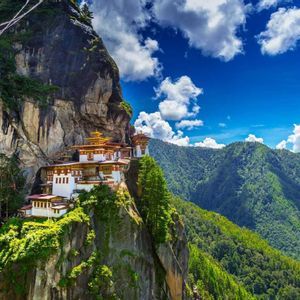 Un complesso monastico con pareti bianche e tetti dorati è costruito in una scogliera a picco che domina una valle di verdi montagne boscose.