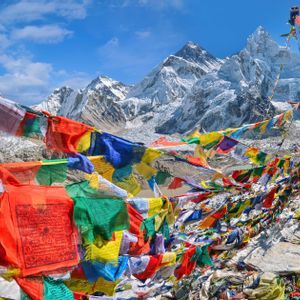 Bandiere di preghiera colorate si estendevano su un punto panoramico roccioso, con montagne innevate e un ghiacciaio sullo sfondo sotto un cielo blu.
