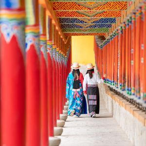 Due donne in abiti tradizionali e cappelli di paglia camminano lungo un corridoio fiancheggiato da pilastri rossi e ruote di preghiera.