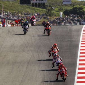 Un gruppo di motociclisti scende a tutta velocità una curva ripida su una pista professionale durante una competizione, con spettatori sullo sfondo.