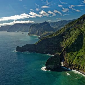 Ripide scogliere verdi si tuffano nell'oceano blu lungo una costa frastagliata sotto un cielo soleggiato con nuvole bianche.