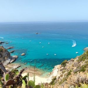Vista dall'alto di una costa rocciosa con una spiaggia appartata e acqua turchese, dove diverse barche e una grande barca a vela fluttuano sotto un cielo limpido.