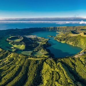 Veduta aerea di una grande caldera vulcanica contenente un lago azzurro, circondata da rigogliose colline verdi, con l'oceano visibile all'orizzonte.