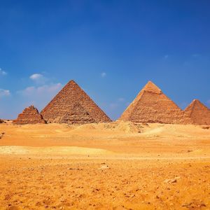 Diverse grandi piramidi si ergono dal deserto dorato sotto un cielo azzurro, con due persone su un cammello e un cavallo in primo piano.