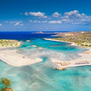 Aerial view of a sandy peninsula stretching into shallow turquoise water, with mountains on the distant coastline under a blue sky.