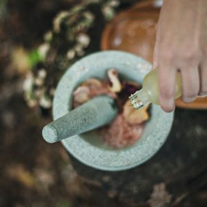 Una vista dall'alto di una mano che versa un liquido da una bottiglia in un mortaio e pestello di pietra con ingredienti naturali all'interno.