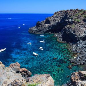 Una vista aerea di diverse barche che galleggiano su acqua limpida e turchese all'interno di una caletta rocciosa, sotto un cielo azzurro brillante.