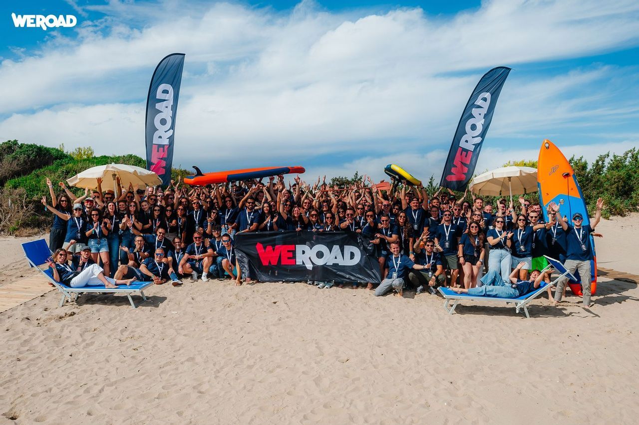 Un grande gruppo WeRoad in posa per una foto su una spiaggia sabbiosa, con striscioni e tavole da surf sotto un cielo blu.
