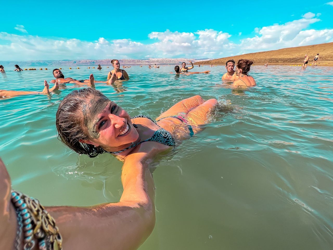 Una donna con del fango sul viso sorride mentre galleggia nell'acqua, sorretta da una mano, durante un viaggio di gruppo WeRoad.