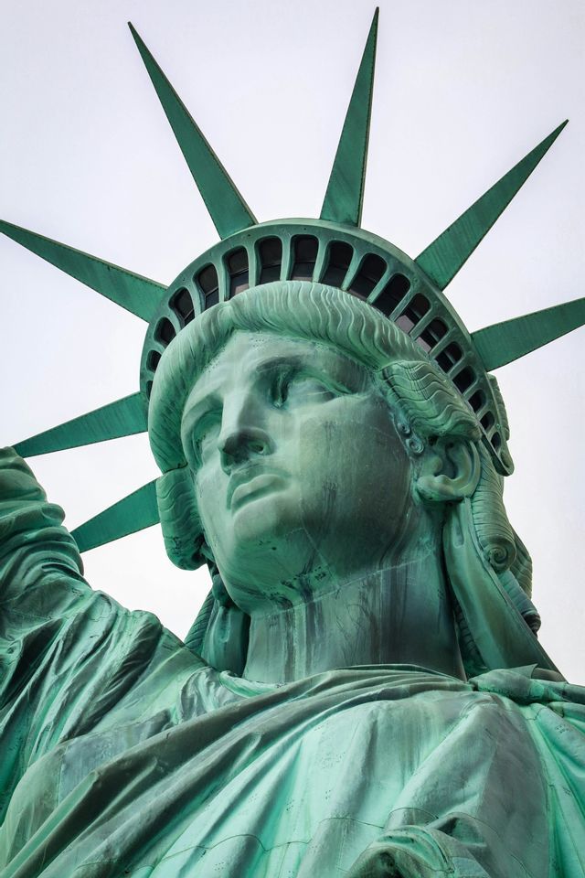 A close-up shot of the Statue of Liberty's face and crown, viewed from a low angle against a white sky.