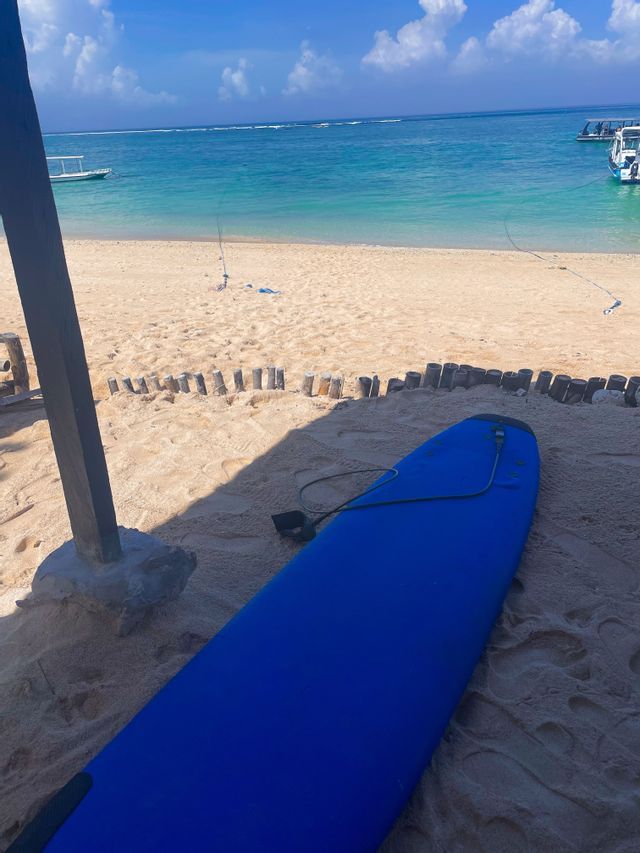 Una tavola da surf blu giace su una spiaggia sabbiosa, con vista sull'oceano turchese e barche sotto un cielo limpido.