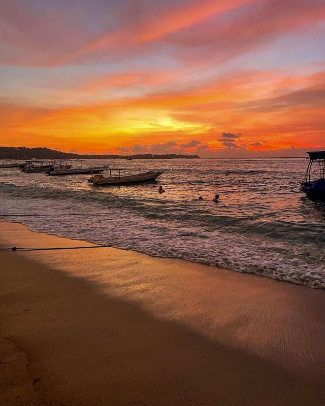 Una spiaggia sabbiosa al tramonto con onde leggere, persone che nuotano nell'oceano e barche ancorate al largo sotto un cielo colorato.