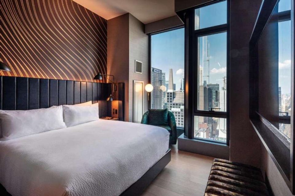 Una camera da letto d'hotel moderna con un letto rifatto e una finestra a tutta altezza che offre una vista sullo skyline di una città.