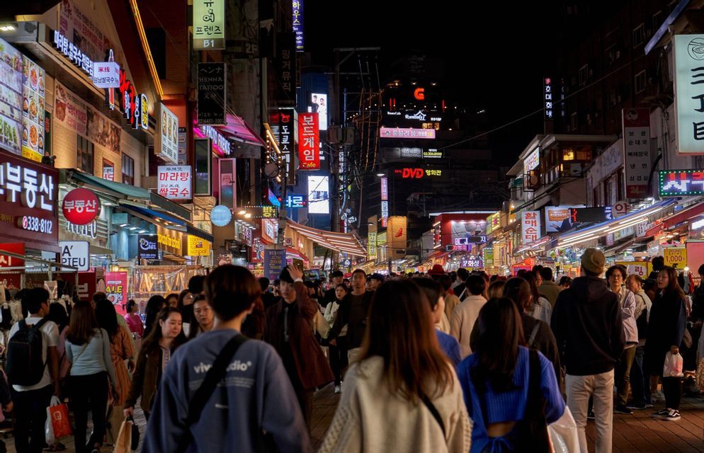 Una grande folla di persone cammina lungo una trafficata via cittadina di notte, illuminata da vivaci insegne al neon con caratteri coreani.
