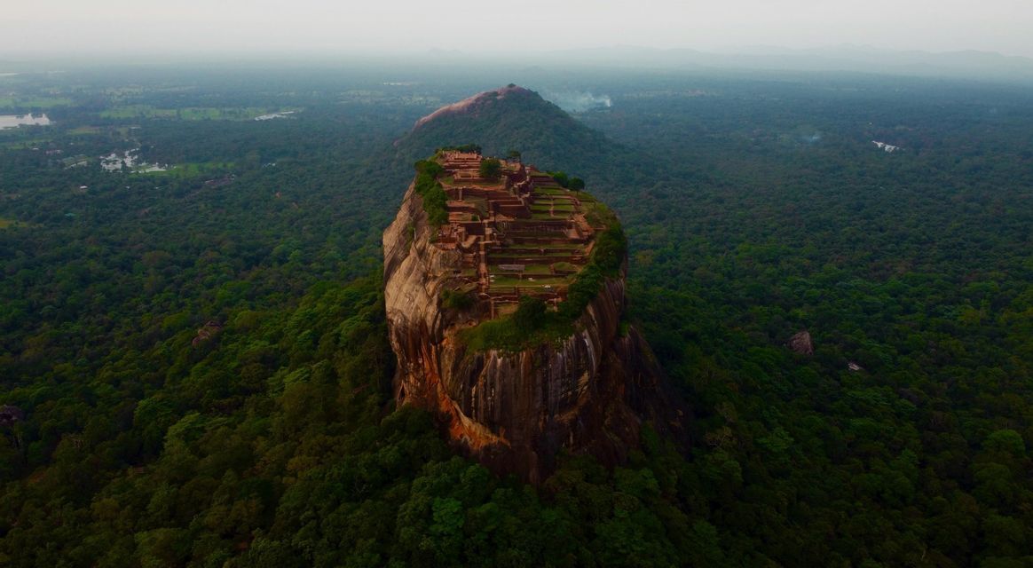 Un'antica fortezza con rovine terrazzate sorge sulla cima di un massiccio monolite roccioso circondato da una fitta giungla verde.