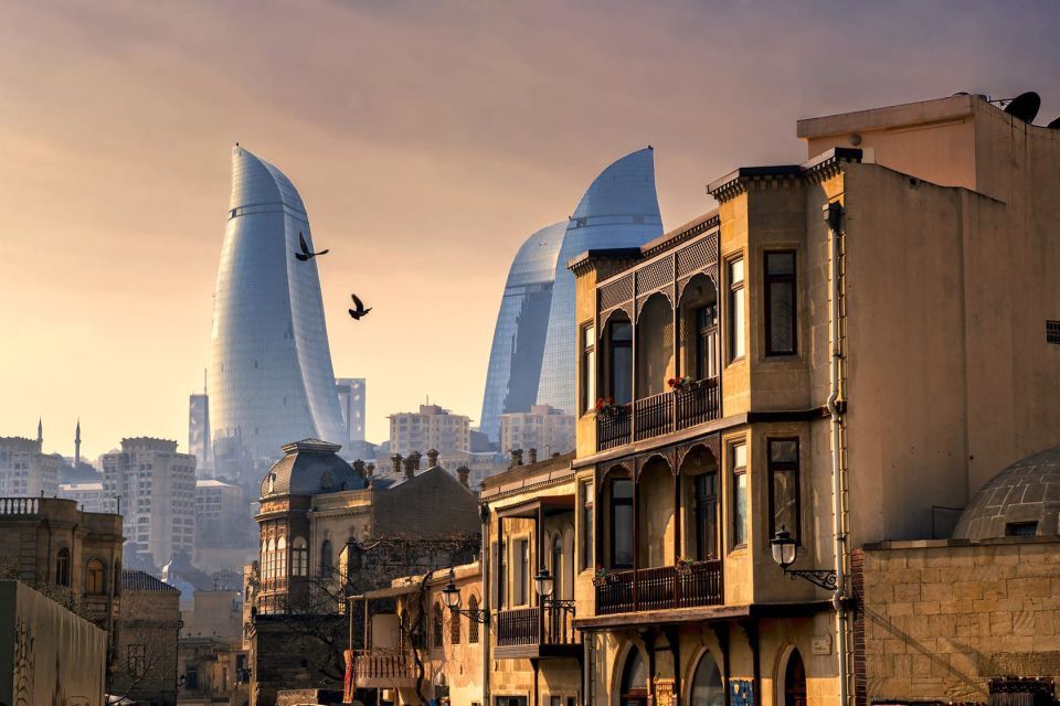 Uno skyline cittadino giustappone edifici tradizionali in pietra con moderni grattacieli di vetro curvi sotto un cielo sfumato al tramonto.