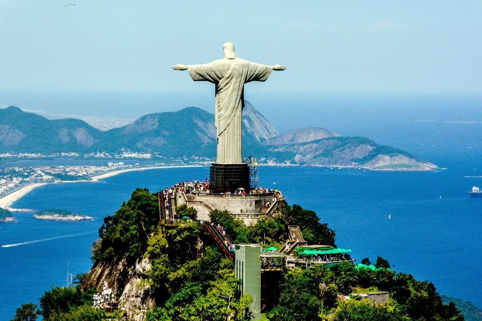 Un'imponente statua in pietra con le braccia aperte si erge su una cima di montagna, affacciandosi su una costa, una città e l'oceano, con turisti alla sua base.