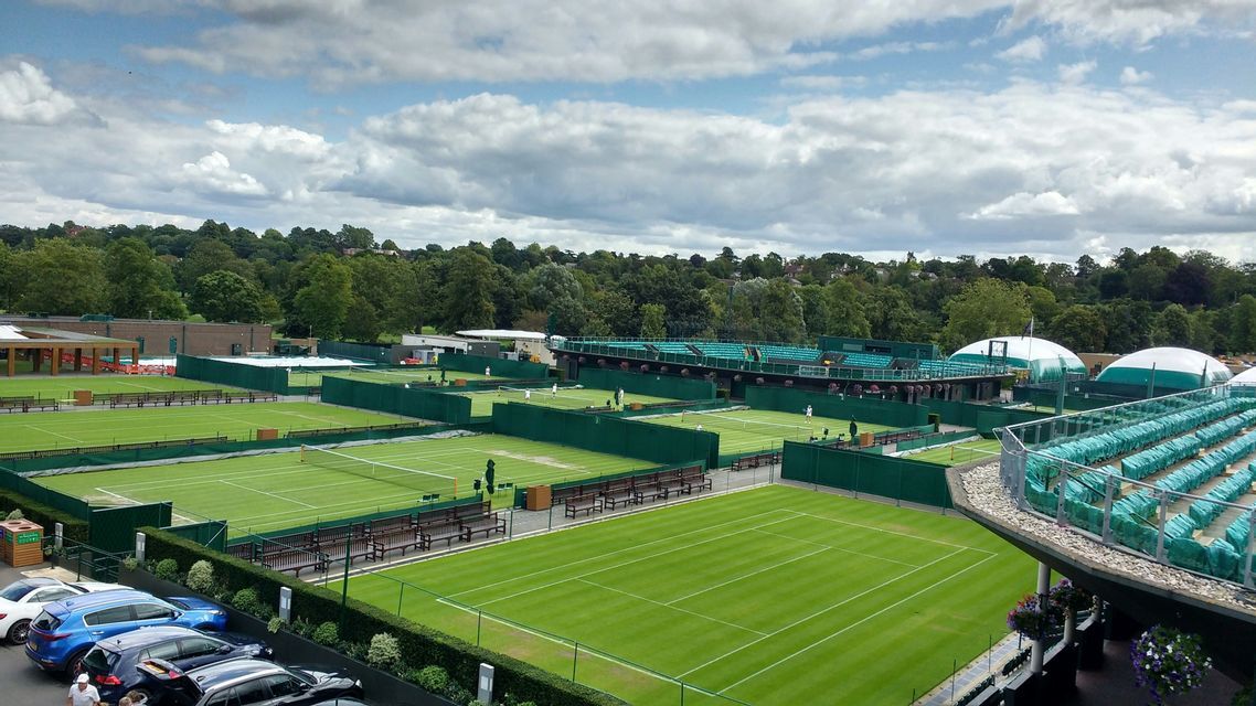 Una vista dall'alto di numerosi campi da tennis in erba verde con persone che giocano, circondati da alberi sotto un cielo nuvoloso.