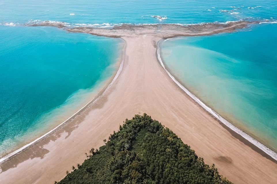 Una vista aérea de una formación de playa con forma de cola de ballena, con agua turquesa a ambos lados y un bosque verde en su base.