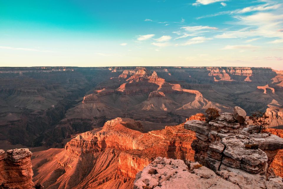 A vast, layered canyon with red rock formations illuminated by golden hour light under a blue sky with clouds.