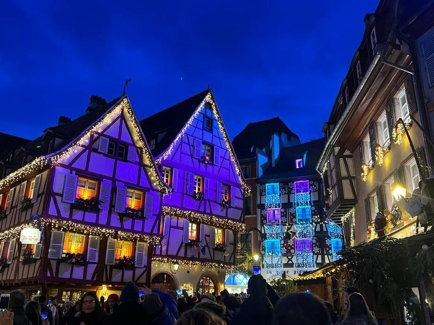 Edificios de entramado de madera iluminados con luces navideñas moradas y blancas sobre una calle concurrida por la noche