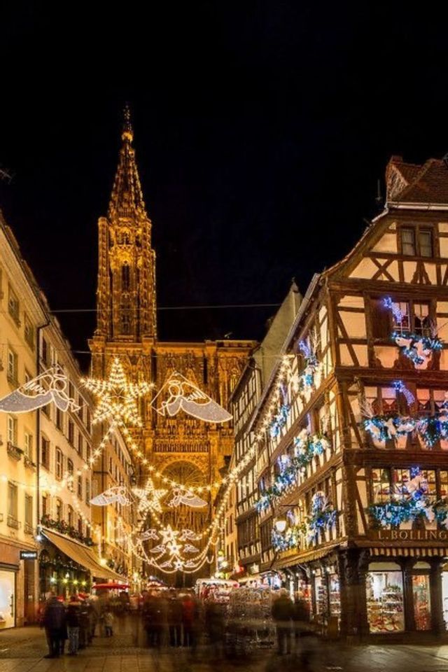 Una calle estrecha con edificios de entramado de madera está decorada con luces de Navidad por la noche, con una alta catedral al fondo.