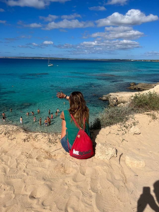 Una mujer con una mochila roja de WeRoad se sienta en un acantilado arenoso, observando a un grupo de viaje nadar en el mar turquesa.