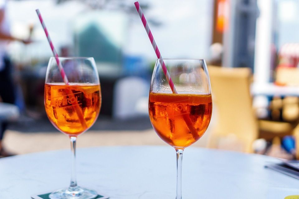 Due bicchieri di Aperol Spritz con cannucce a strisce rosa e bianche su un tavolo bianco in un ambiente esterno.