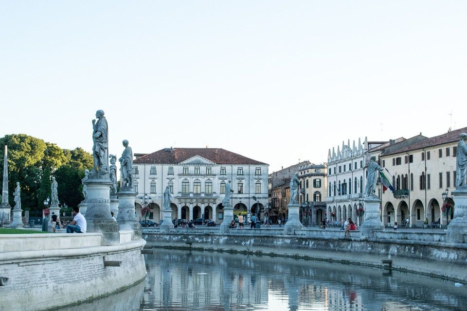 Un canale ellittico fiancheggiato da numerose statue in pietra su piedistalli, con edifici classici e alberi sullo sfondo.