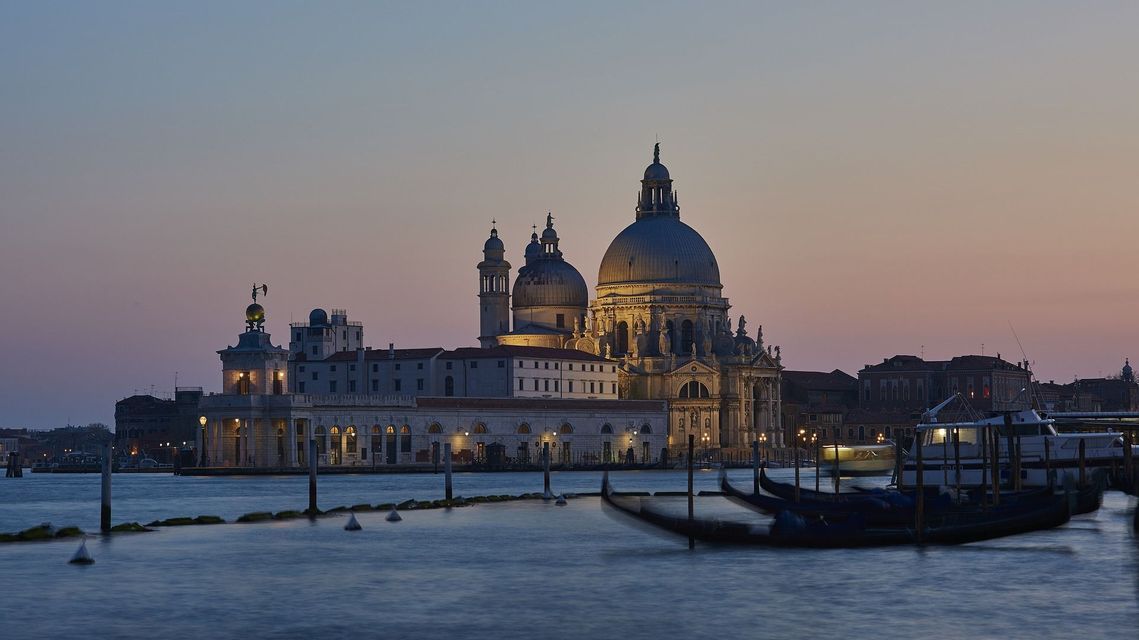 Una basilica illuminata con grandi cupole sul lungomare al tramonto, con gondole ormeggiate in primo piano.