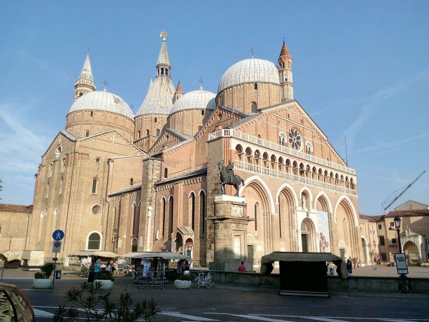 Una grande cattedrale in mattoni con cupole multiple si erge dietro una piazza con una statua equestre e bancarelle del mercato sotto un cielo blu.