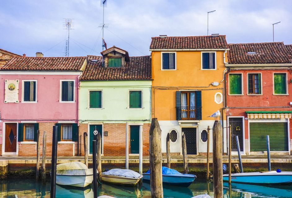 Una fila di edifici colorati con tetti di tegole costeggia un canale dove diverse piccole imbarcazioni sono ormeggiate a pali di legno.