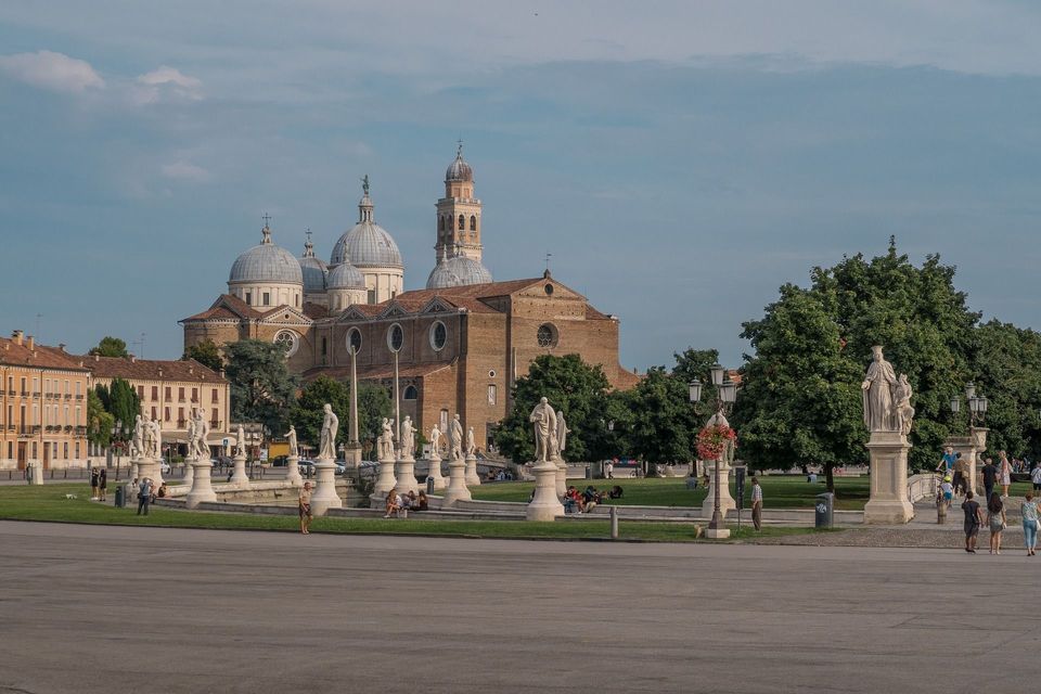 Una grande basilica in mattoni, con cupole multiple, si erge dietro una piazza pubblica che presenta un prato verde circondato da numerose statue bianche.