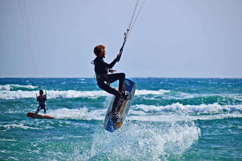 Un kitesurfer in muta nera è sospeso a mezz'aria su onde oceaniche blu mosse sotto un cielo sereno.