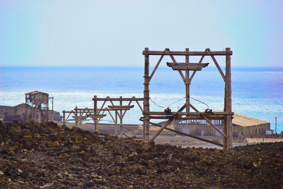 Strutture industriali in legno fatiscenti su una costa rocciosa con il mare blu sullo sfondo