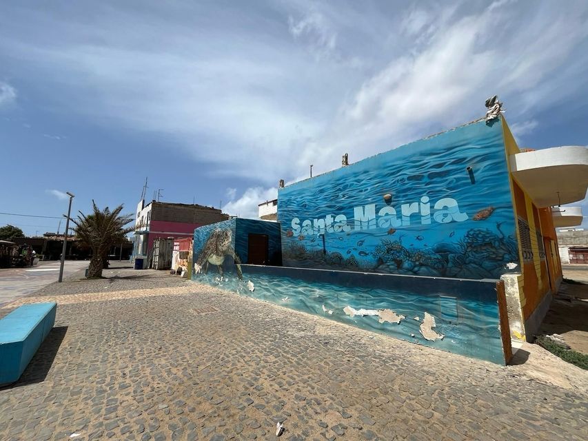 Un grande murale blu raffigurante una scena sottomarina con una tartaruga marina e le parole 'Santa Maria' su un edificio accanto a una strada acciottolata.