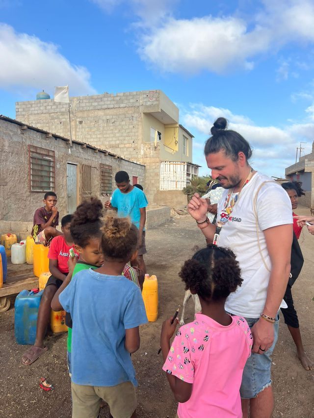 Un uomo in viaggio di gruppo con WeRoad, con il man-bun, sorride interagendo con un gruppo di bambini locali in una strada di paese.