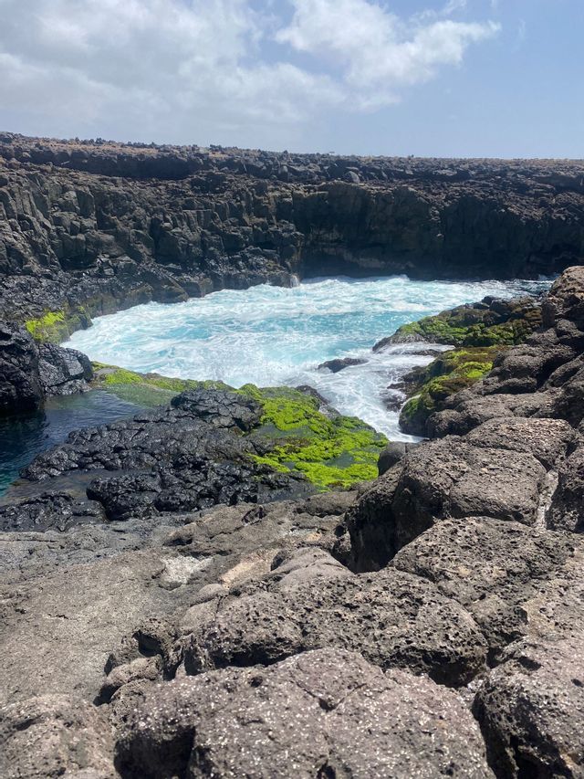 Una piscina naturale di acqua turchese ribollente è circondata da scure rocce vulcaniche con chiazze di muschio verde brillante.