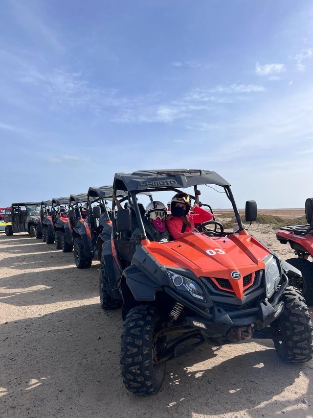 Un viaggio di gruppo WeRoad su una lunga fila di dune buggy arancioni parcheggiati su una strada sabbiosa sotto un cielo azzurro.