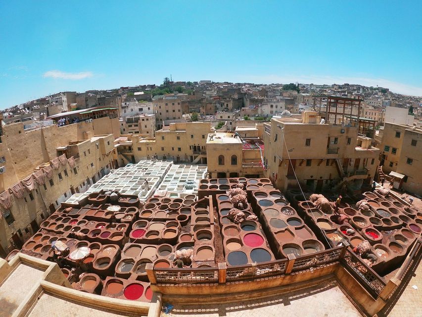 A high-angle fisheye view of a traditional tannery with numerous stone vats containing colorful dyes, set amidst a dense cityscape.