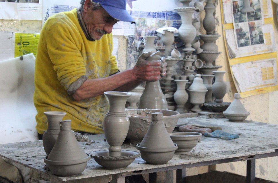 An artisan in a yellow sweater and blue cap molds a clay vase on a pottery wheel, surrounded by finished pottery in a workshop.