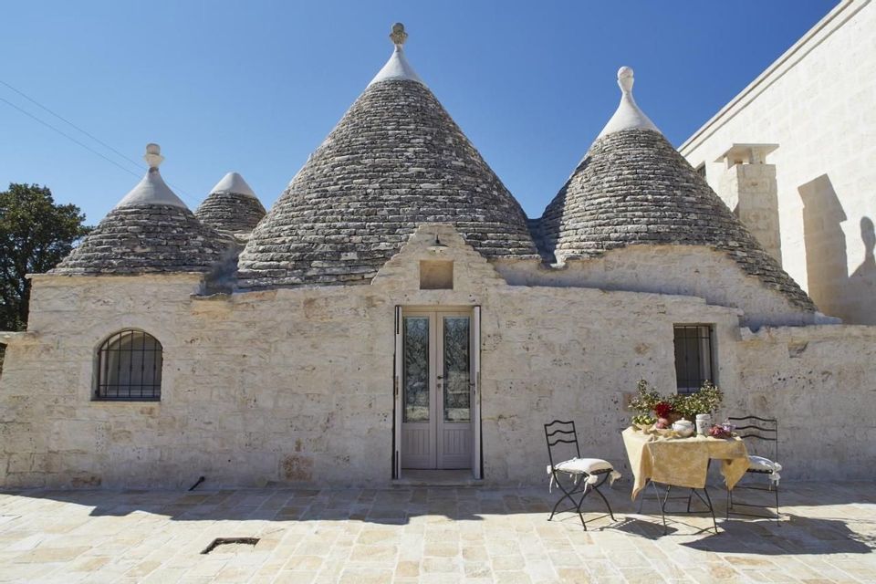 Un trullo tradizionale con tetti conici in pietra sotto un cielo azzurro limpido, con un tavolo e due sedie disposti sul patio in pietra.